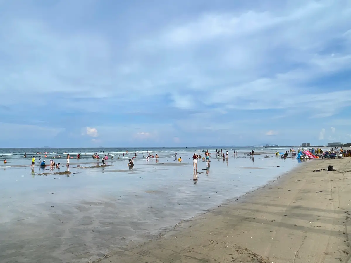 Wide shallow sea with many people wading along a long sand beach under a cloudy sky