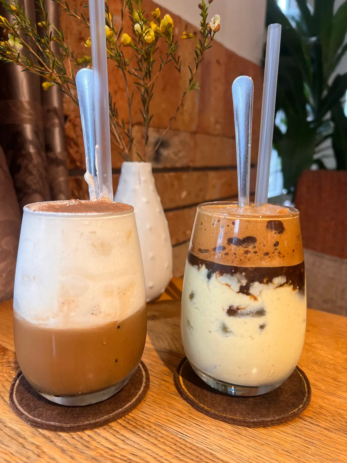 Two tall glasses of iced coffee with cream and chocolate layers beside a vase