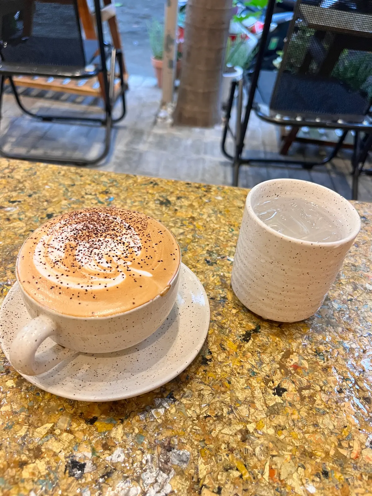 Cappuccino with cocoa dust on a speckled saucer beside a cup of water on a gold terrazzo table