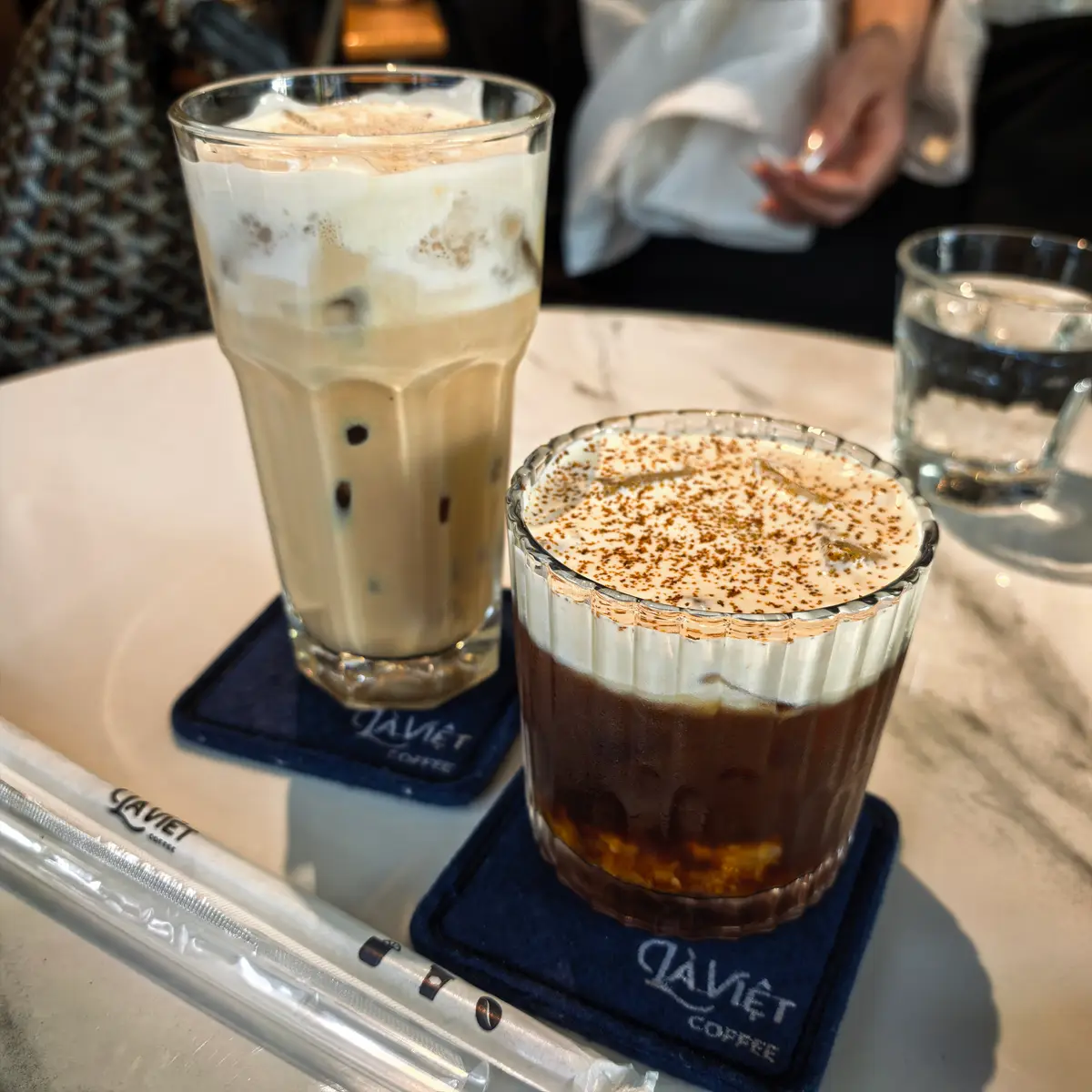 Iced milk coffee and a glass of coffee topped with foam on a marble table