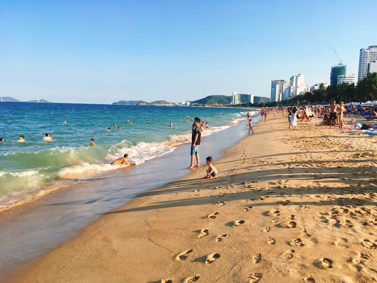 Visitors walking and paddling on the sand at Nha Trang beach on a clear day, with the city skyline in the distance