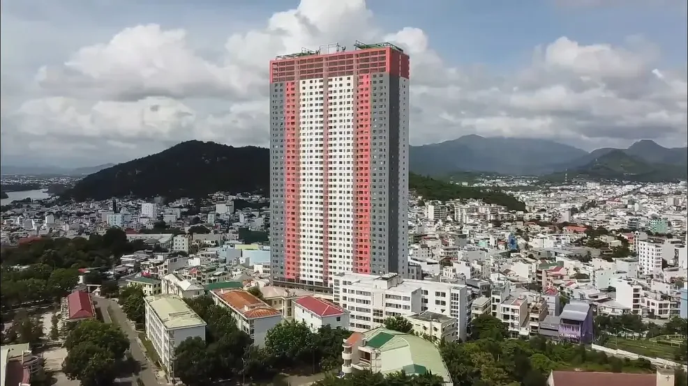 Tall red and white apartment tower rising above low rooftops and green hills in Nha Trang