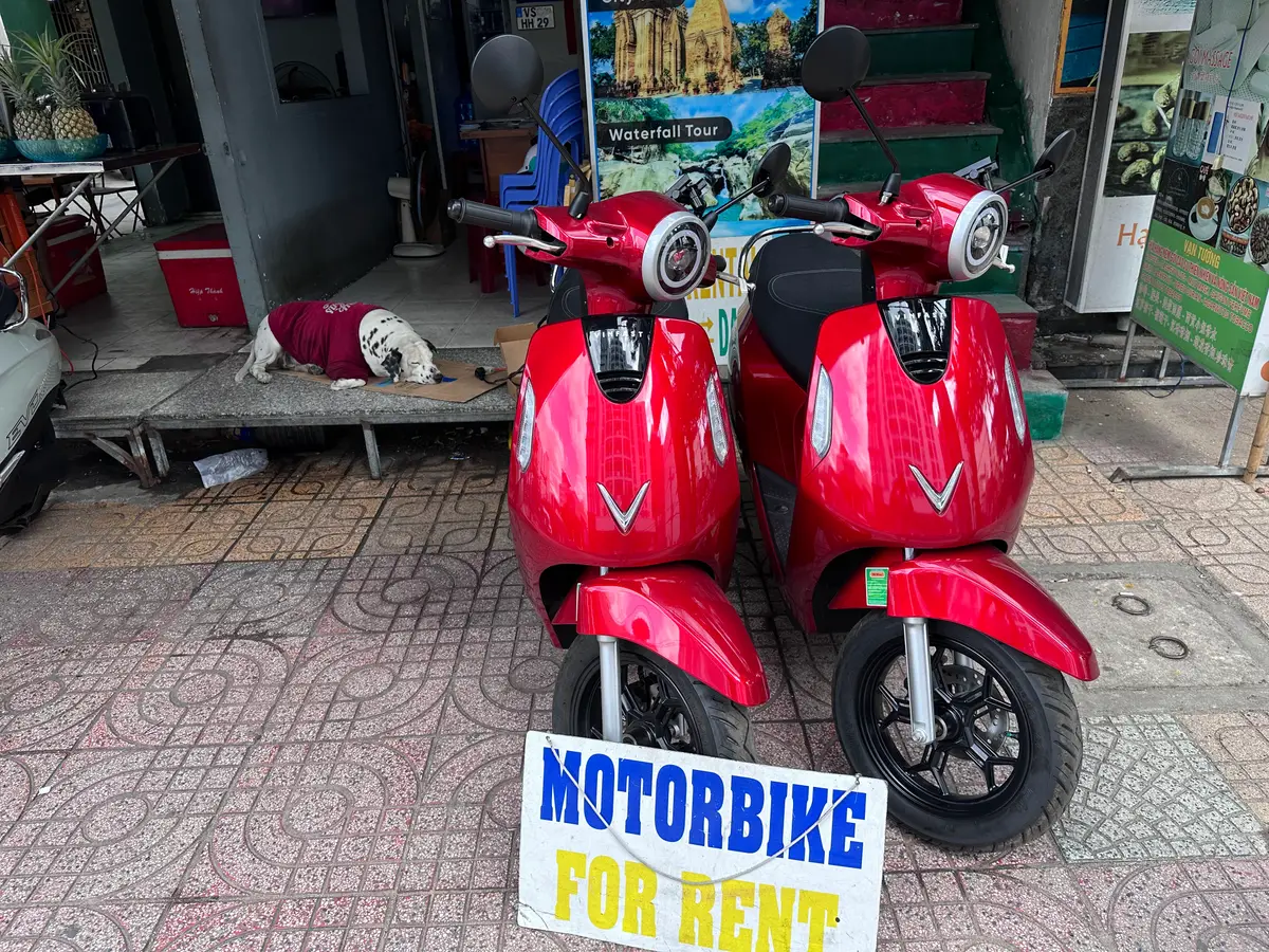 Two red electric scooters outside a shop beside a Motorbike For Rent sign