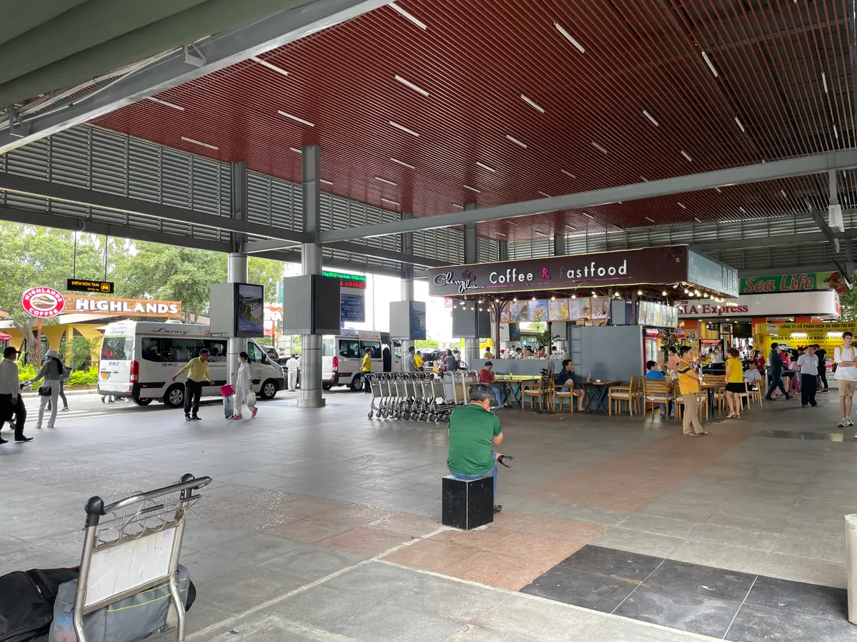 Covered forecourt outside Cam Ranh International Airport with a minibus, a luggage trolley and a row of kiosks under a red ceiling