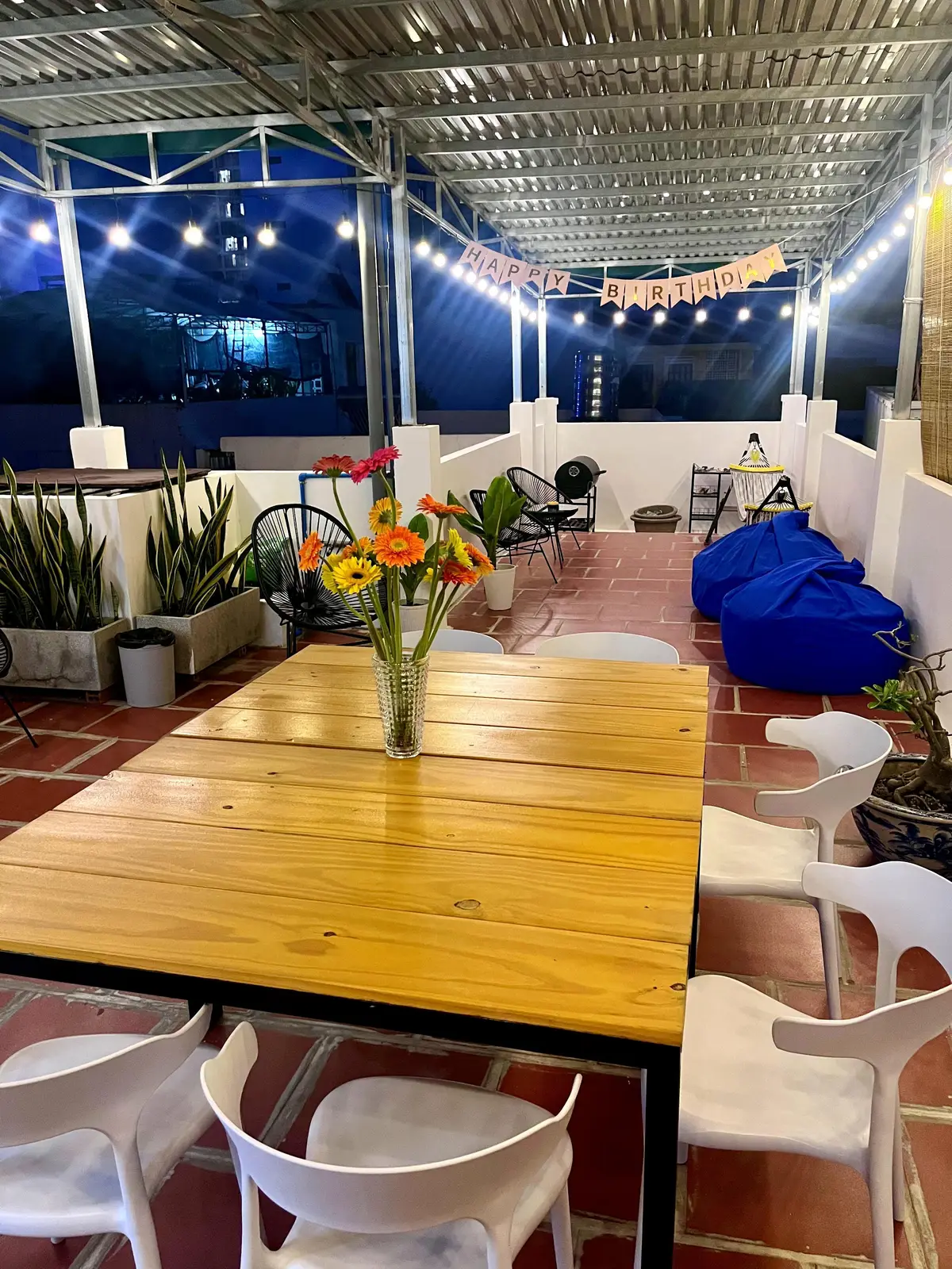 Sunny terrace with a long wooden table, white chairs, blue bean bags and potted plants on red tiles