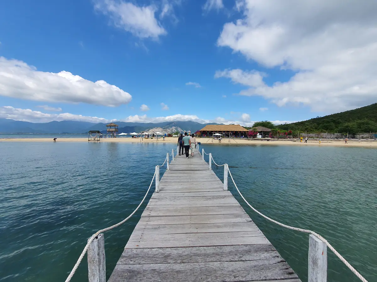 Wooden pier with rope railings leading toward a beach hut and green hills