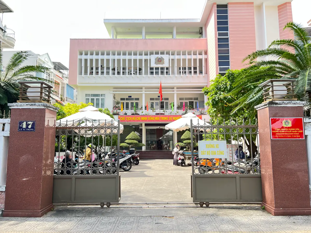 Entrance gate of the Khanh Hoa immigration office with a red sign on the gate pillar, and a pink building behind palm trees and parked motorbikes.