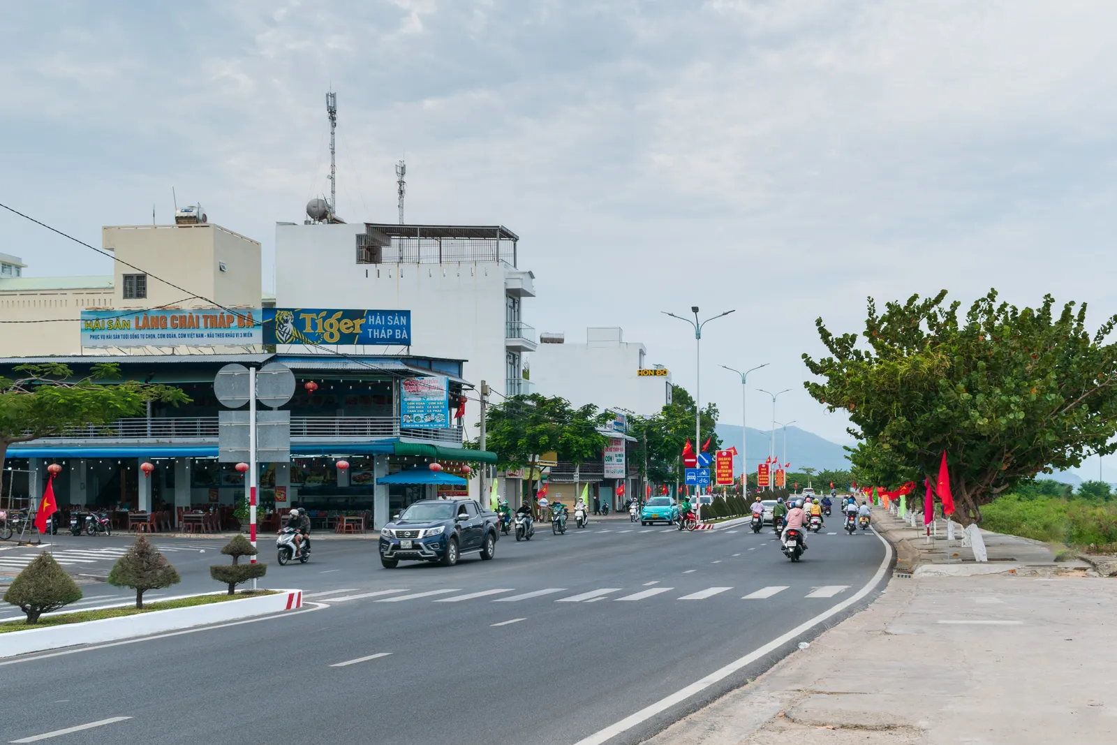 A wide road in Nha Trang with painted crossings, motorbikes, shopfronts with signs on the left and red flags along the kerb.