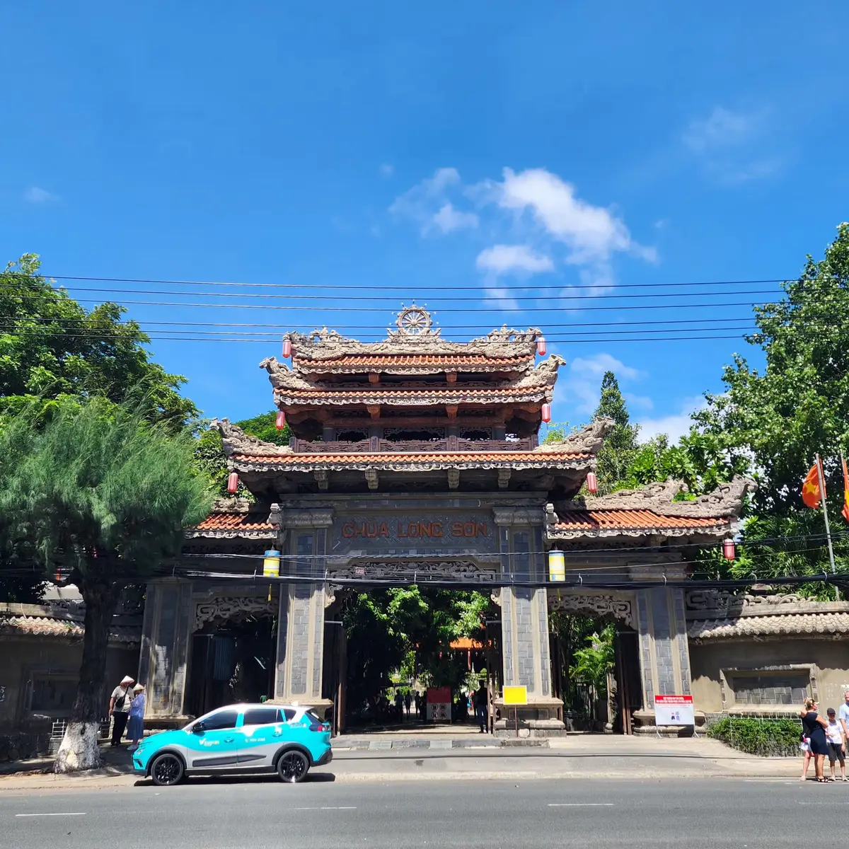 Gate of a pagoda with a tiled triple roof seen from the street, with trees on each side and a car parked in front