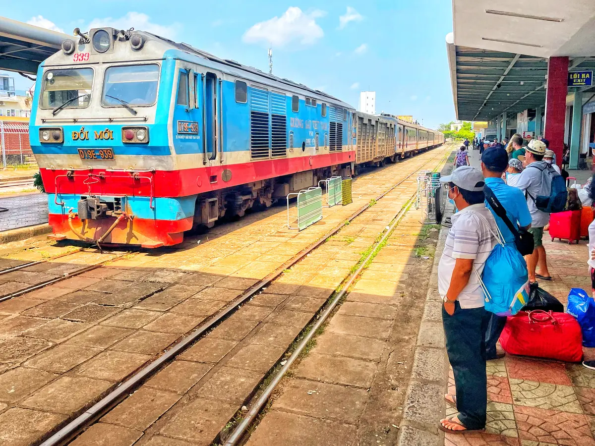 Blue and red passenger train at a platform of Nha Trang railway station with travellers carrying backpacks