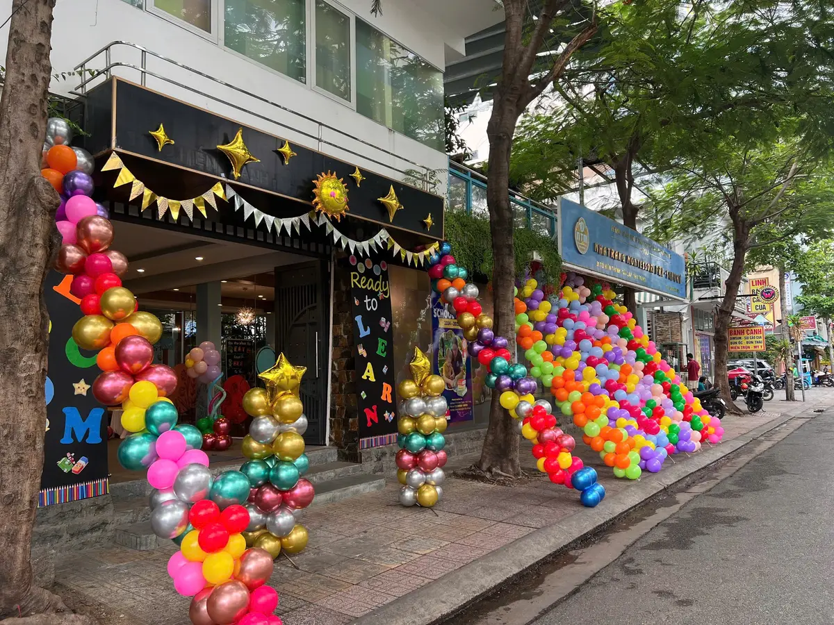 A shopfront decorated with columns of gold, red, green and blue balloons on a tree-lined street.
