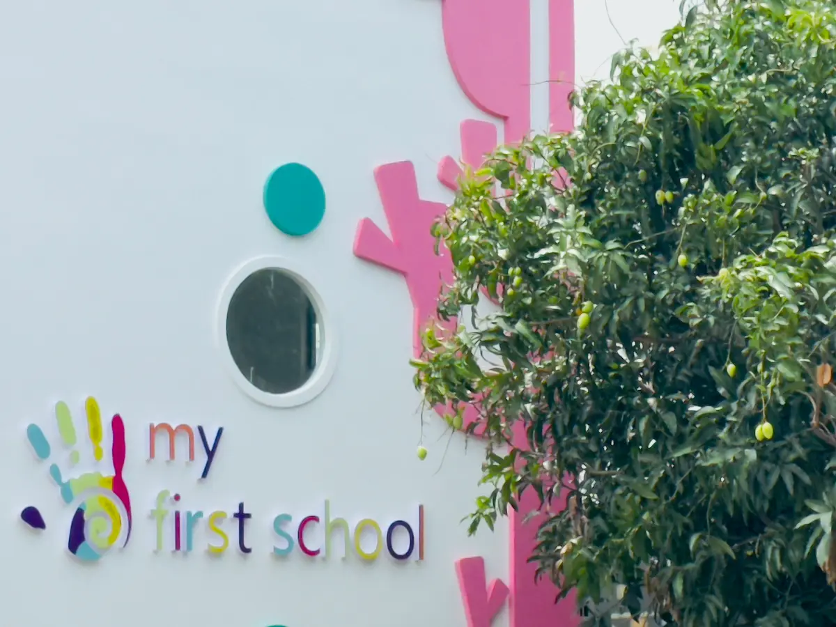 A white wall with a coloured handprint logo reading my first school beside a leafy tree.