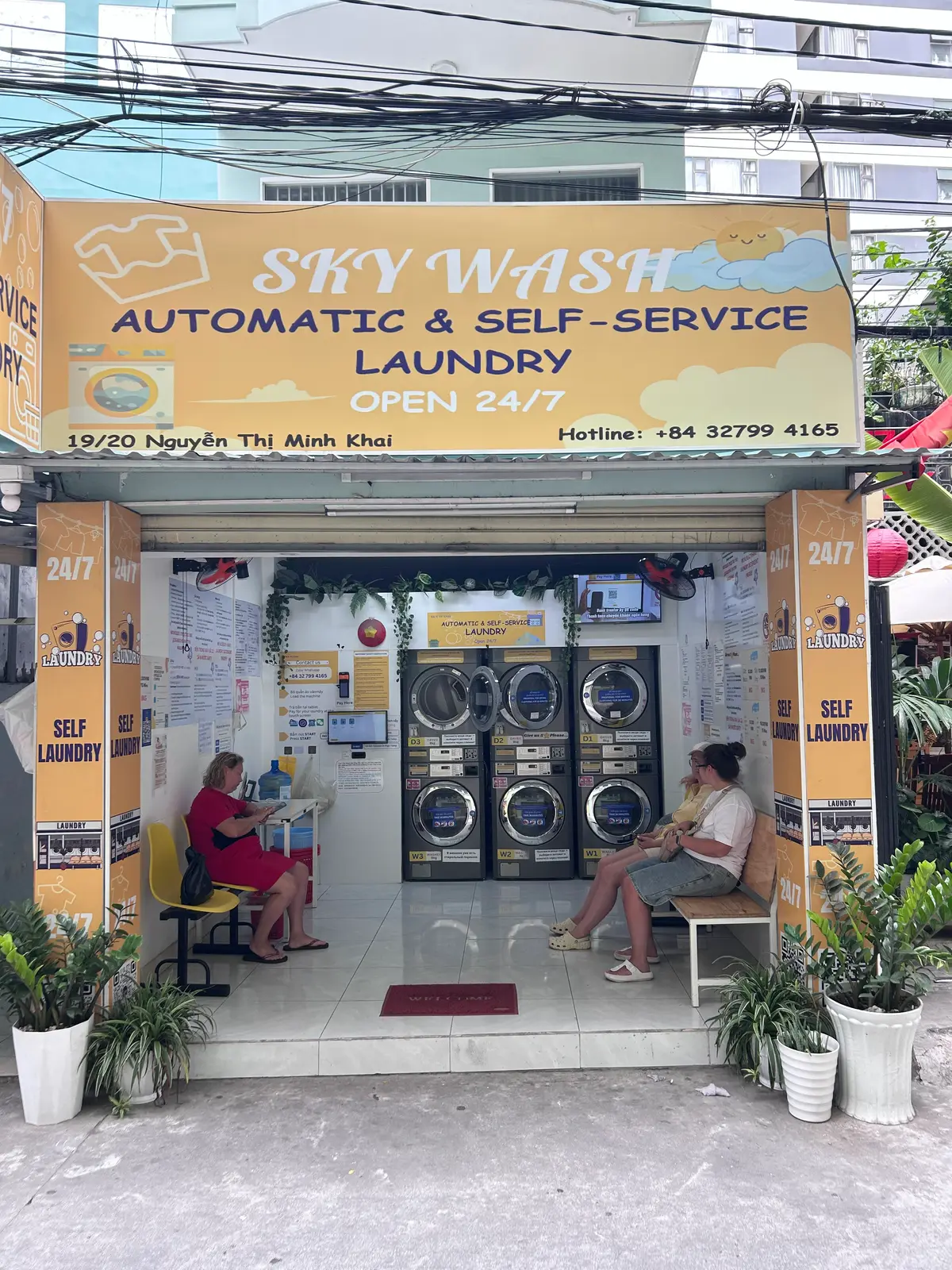 Yellow self-service laundromat storefront with machines and a round-the-clock sign