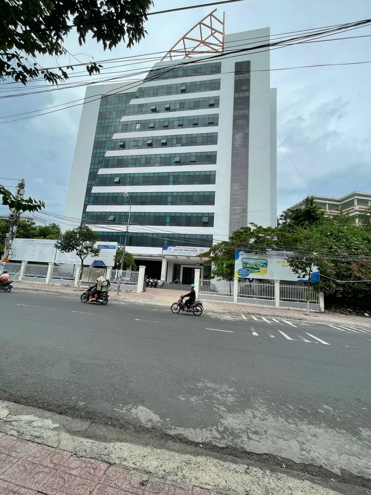 A tall glass-fronted university building behind a fence and a blue sign on a street with motorbikes.