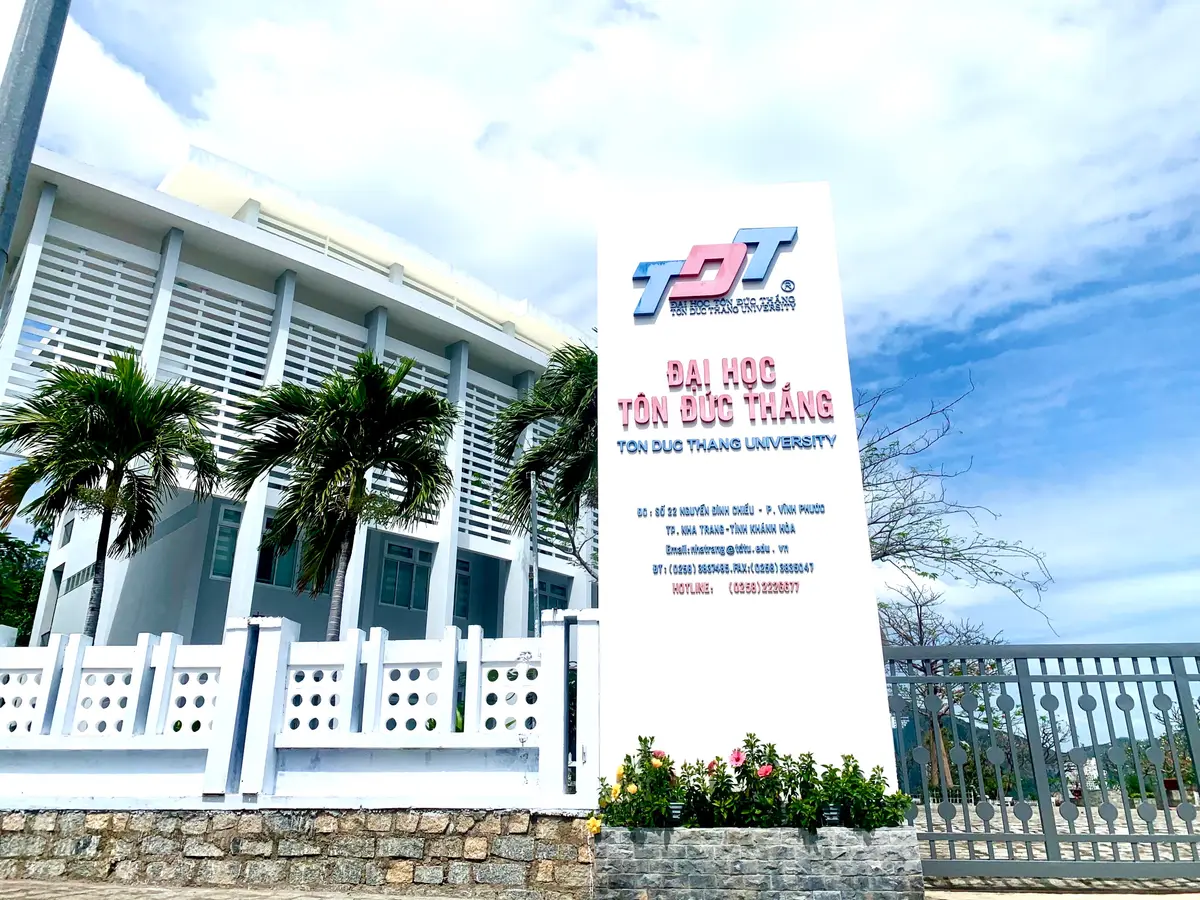 A white stone entrance sign with a red logo and the words Ton Duc Thang University beside a white railing.