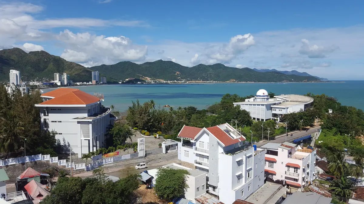 A view over red-roofed buildings and white houses to a calm bay and green mountains.