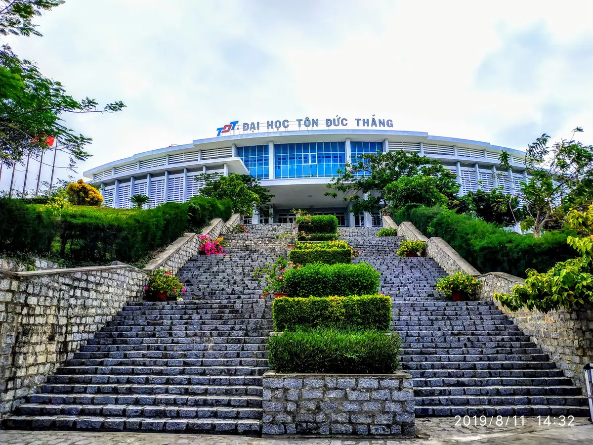 Wide stone stairway with flower beds and clipped hedges leading up to a white university building with blue windows