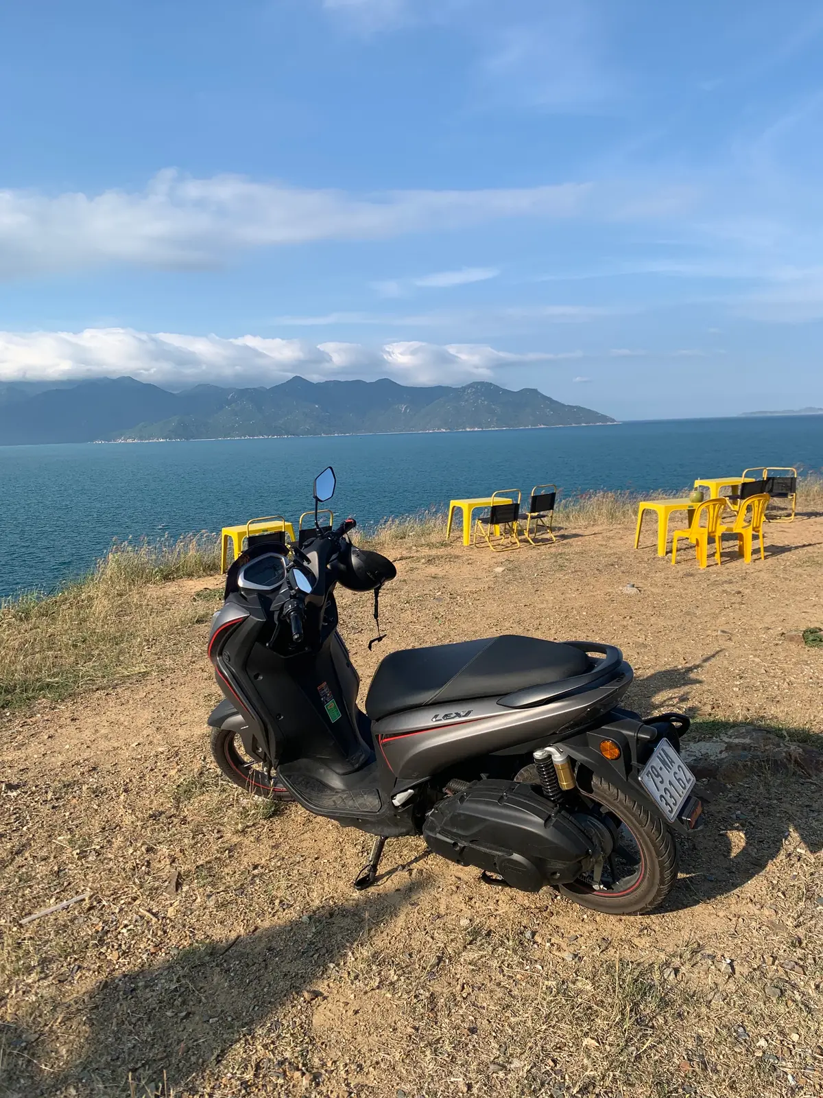 Dark scooter parked on a dirt viewpoint above the sea, with yellow tables in the background