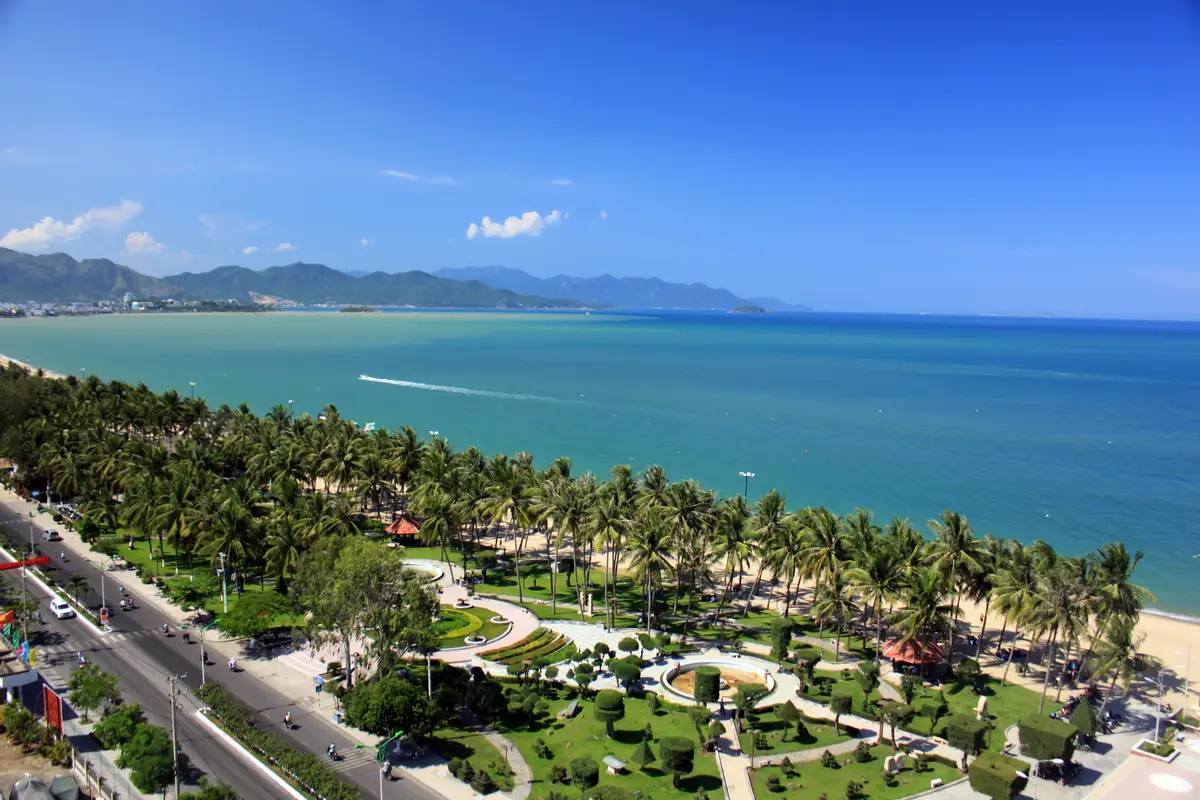 Green seafront park with palm trees and winding paths beside a wide blue sea and distant hills