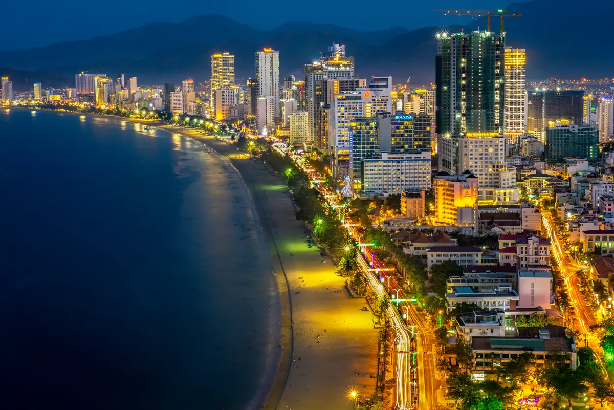 Night view of a curved beach, a lit coast road and glowing hotel towers beside dark water