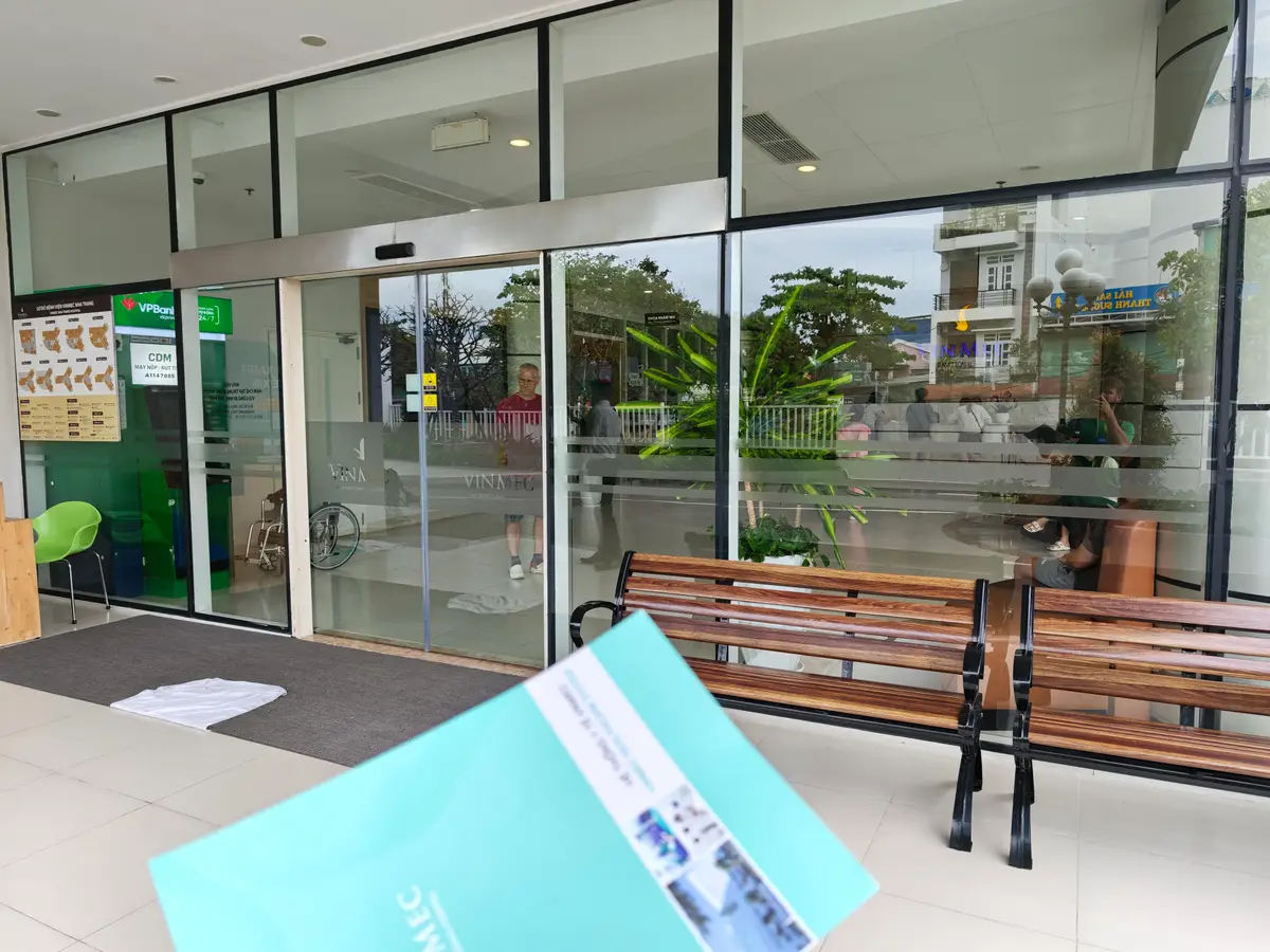 Glass entrance doors of Vinmec Nha Trang hospital with a wooden bench and a leaflet in the foreground