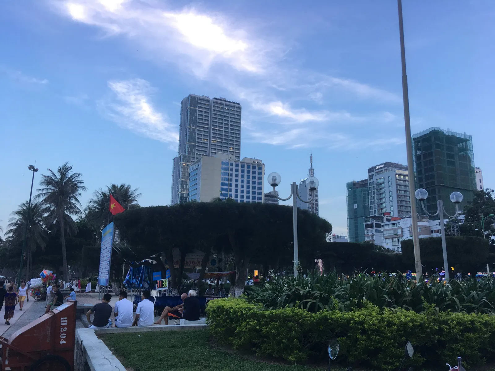 High-rise towers and palm trees beside a seafront park in Nha Trang, with people sitting on the low wall in the foreground.