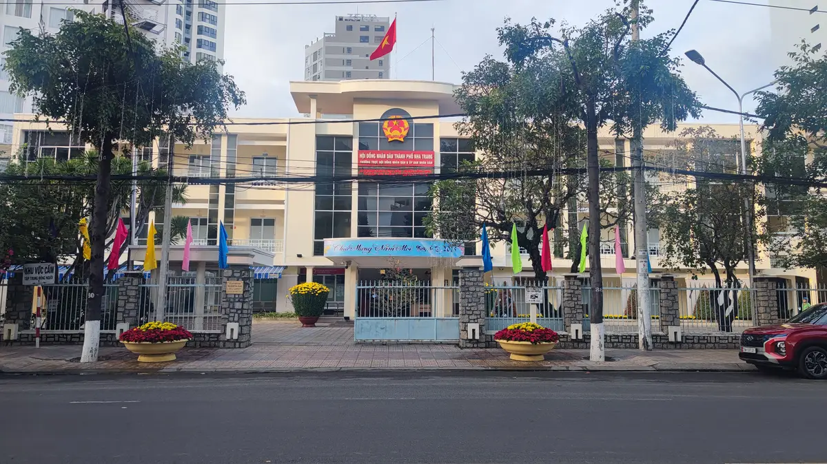 Cream-coloured government building with a national emblem above the entrance, flags along the front and a gate in the foreground