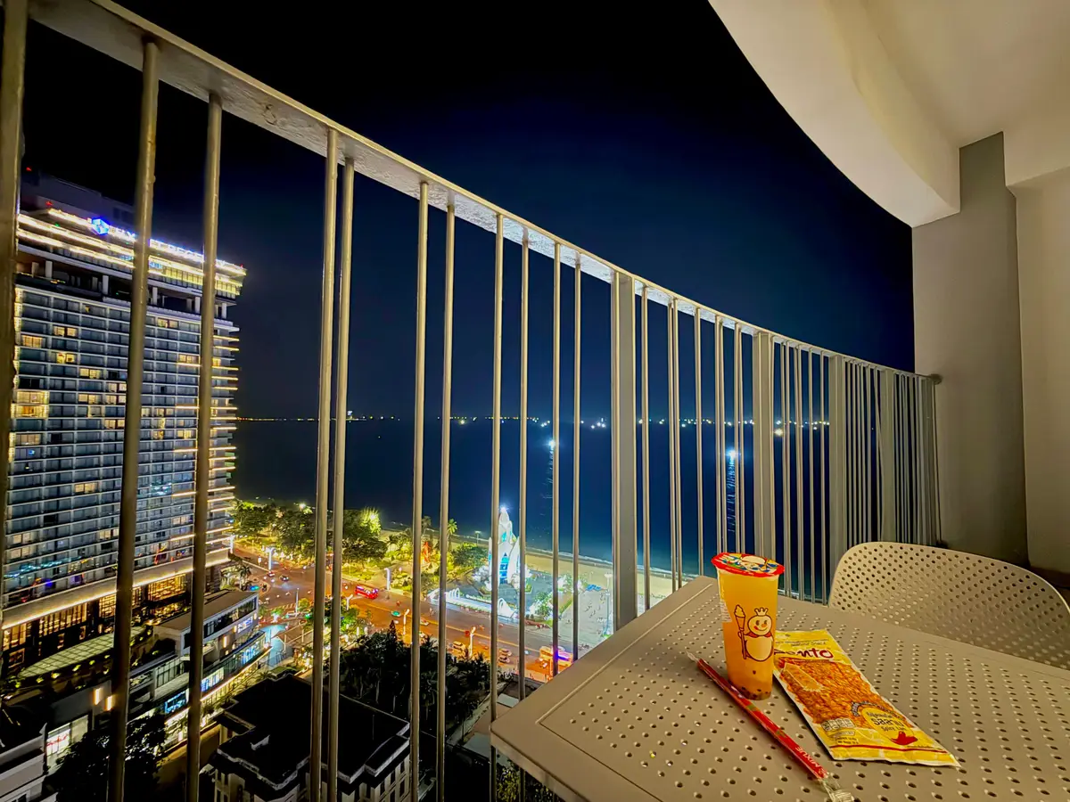 Evening view from a glass-railed balcony with a small table, towers opposite and a lit street below