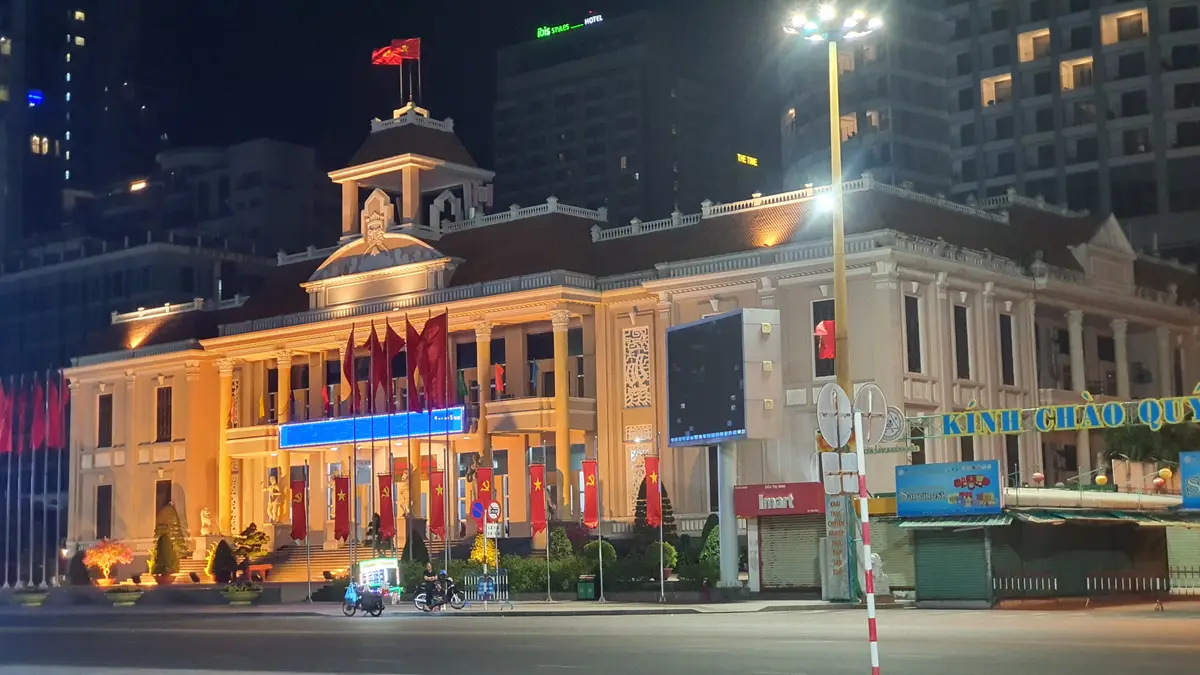 Yellow-lit building with a small tower on top, red flags on the facade and a street with road signs in front, photographed at night