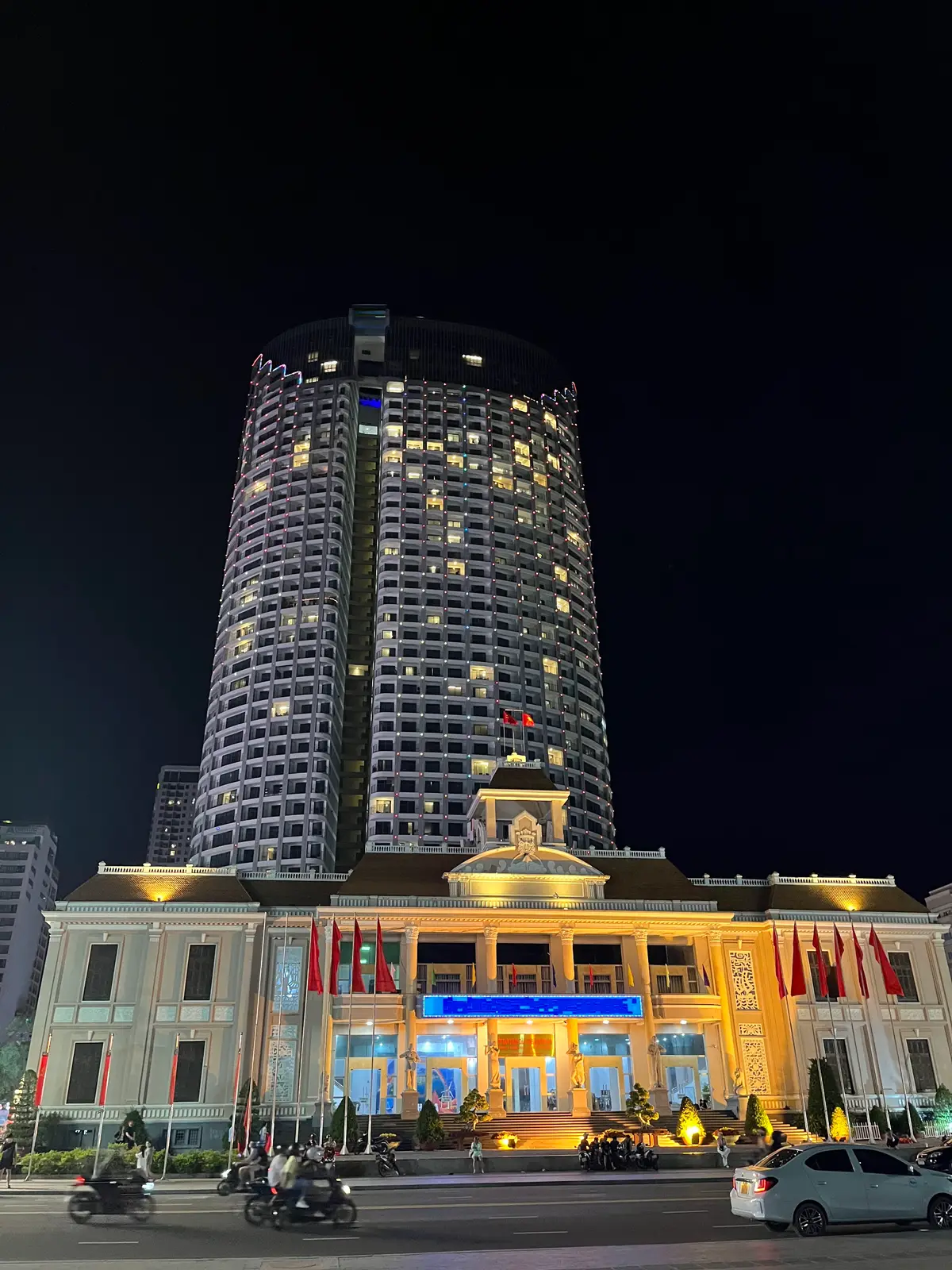 Two lit apartment towers at night rising behind a yellow-lit low building