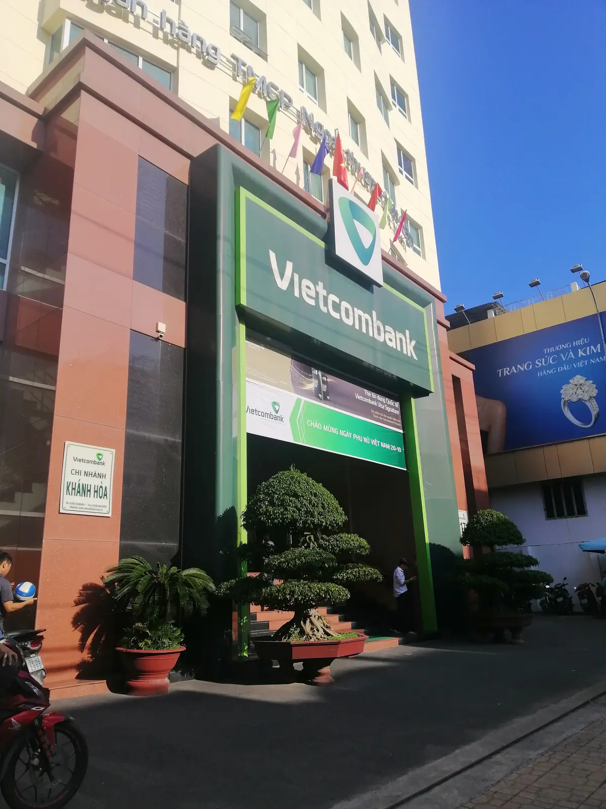 Green bank sign at the entrance of the branch with a small plaque beside it and a trimmed tree in front.