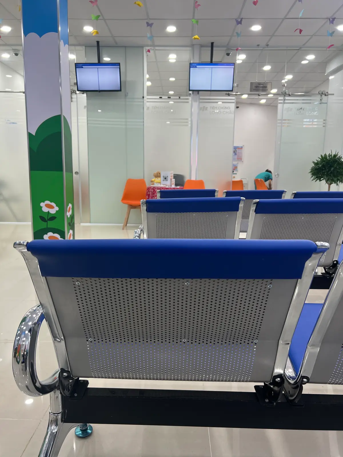 Blue waiting chairs in a row with orange chairs behind them inside a vaccination centre.