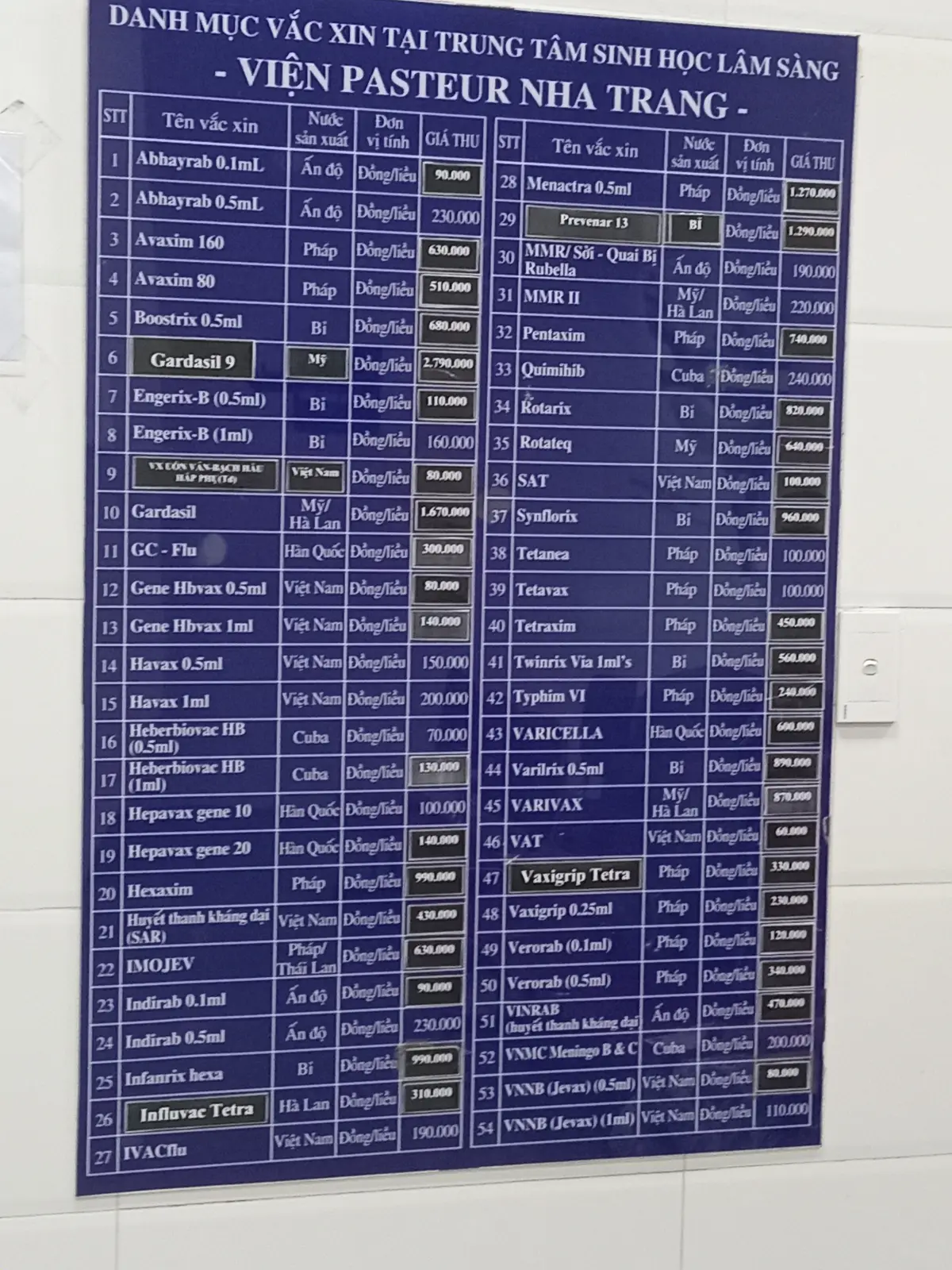 A dark blue price board listing vaccine names, countries and prices in Vietnamese.