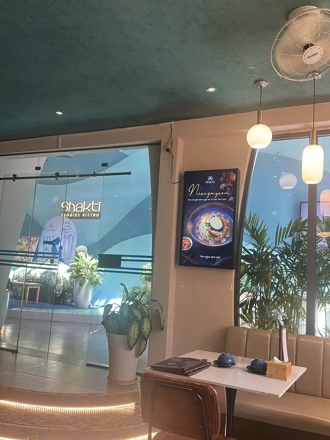 Glass-fronted restaurant with a lit sign and tall indoor plants
