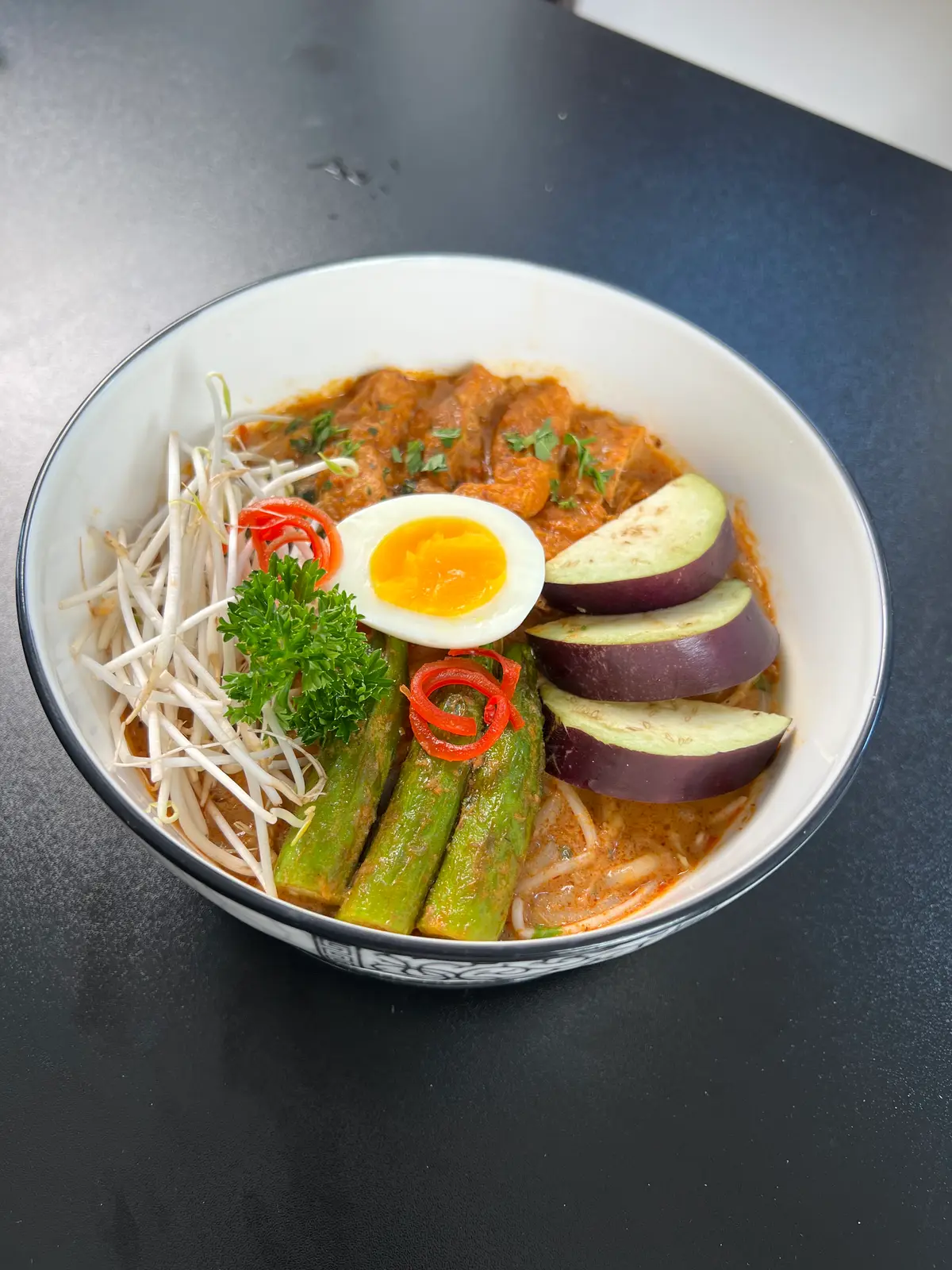 Bowl of noodle soup in red broth topped with a halved egg, aubergine and bean sprouts
