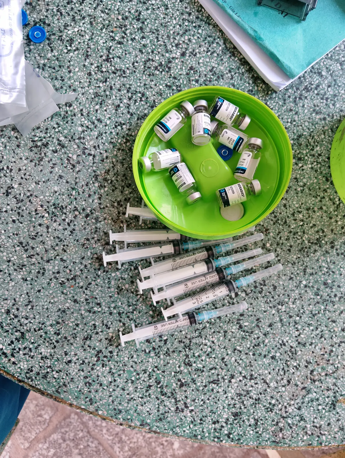 Vaccine vials in a green dish above a row of syringes on a speckled floor