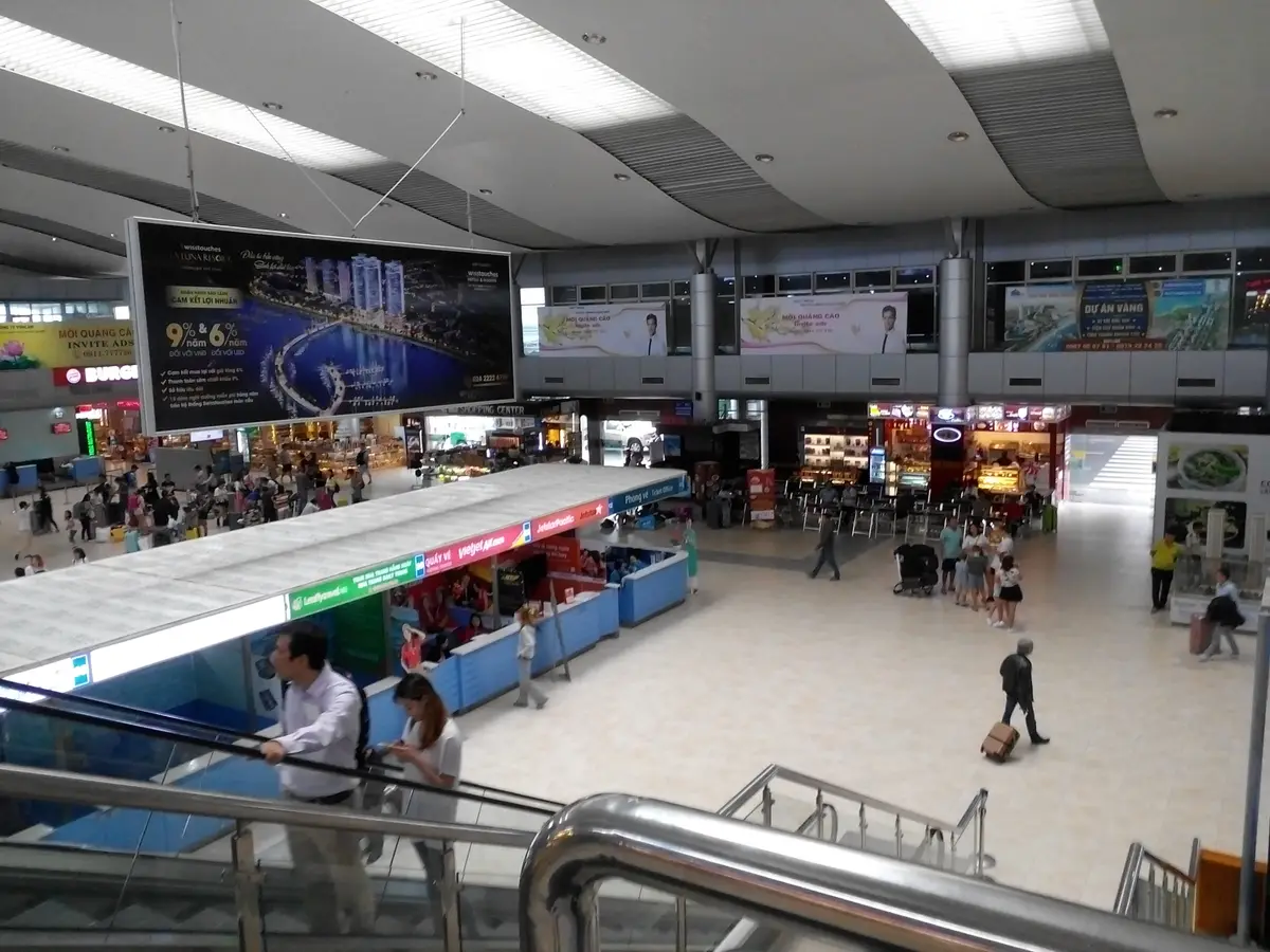 Arrival hall at Cam Ranh airport seen from the escalator, with a long counter, shop signs and travellers walking below.