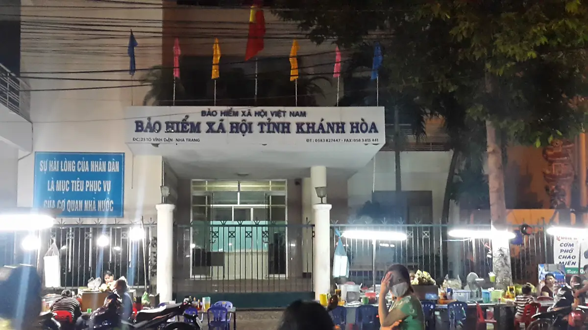 Front entrance of a government office with a sign reading Bao hiem xa hoi tinh Khanh Hoa above the doors and flags along the roofline.