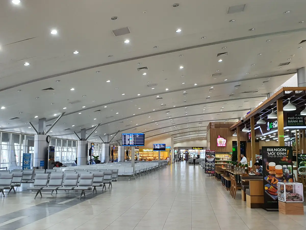 Bright airport hall with rows of seating, a cafe area on the right and a departures board in the distance.