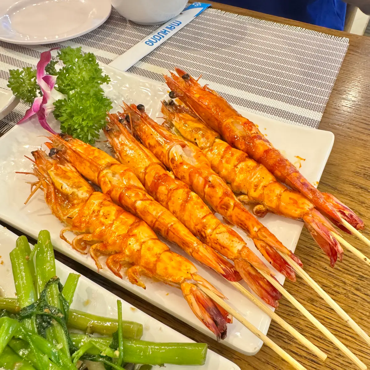 Row of grilled prawns on bamboo skewers beside herbs and a flower garnish