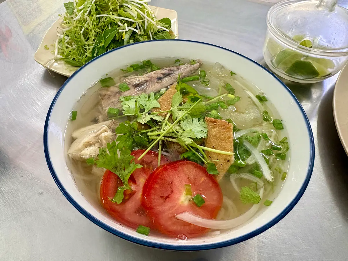 Bowl of fish noodle soup with fish slices, tomato and herbs on a steel table