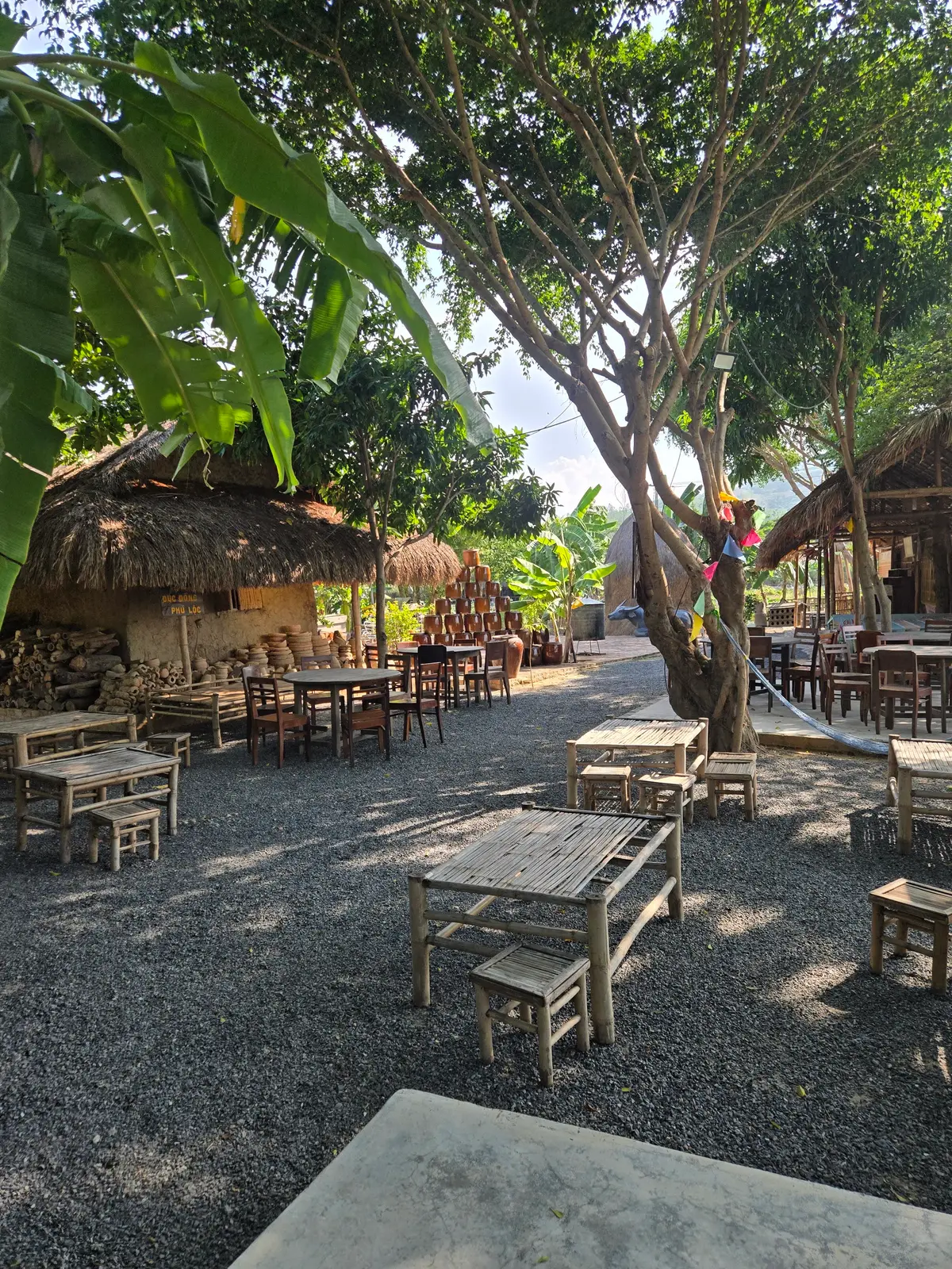 Shaded courtyard with wooden tables and stools under trees and thatched roofs